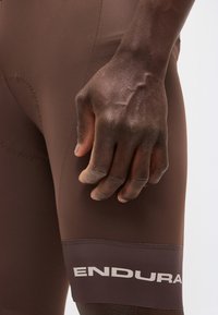 Κοντινό πλάνο ενός σκουρόχρωμου χεριού που ακουμπάει δίπλα σε μηρό με καφέ σορτς ποδηλάτου, με το κείμενο «ENDURA» στη λωρίδα του ποδιού.