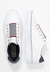 Baskets en toile blanche avec un panneau textile rayé, des œillets dorés et une touche bleu marine au talon. Semelle en caoutchouc texturée.