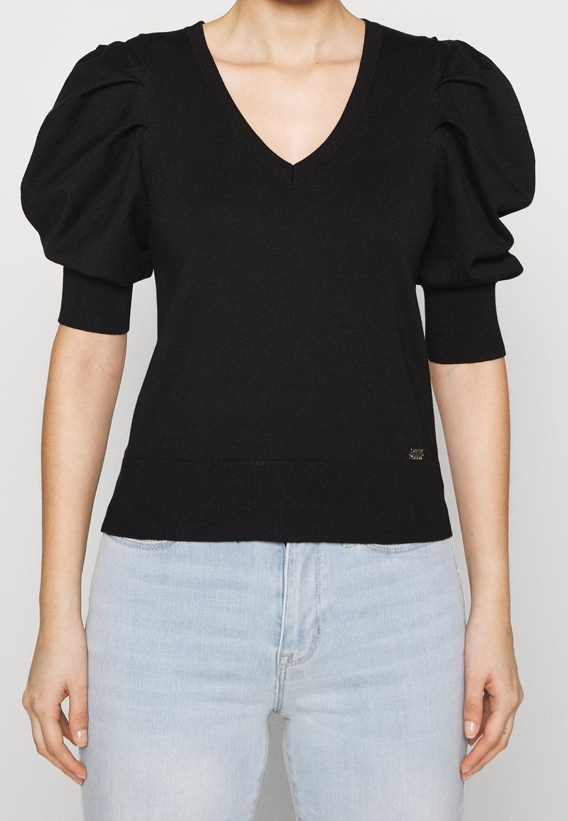 Femme portant un pull noir col en V avec des manches courtes bouffantes et un jean bleu clair, vue des épaules jusqu'aux hanches.