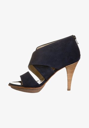 Sandale à talon haut en suede bleu marine avec un design à croix, bout ouvert et fermeture éclair au dos. Dotée d'un talon à texture bois.