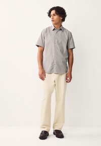 Jeune homme aux cheveux bouclés portant une chemise à manches courtes à motifs, un pantalon crème et des mocassins foncés, debout devant un fond blanc uni.