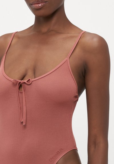 Close-up of a person wearing a ribbed, dusty rose swimsuit with thin straps and a small tied bow at the chest.
