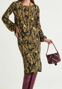 Abito lungo floreale con motivi verdi e beige, maniche lunghe, sfondo nero; borsa bordeaux con design geometrico; stivali bordeaux abbinati.