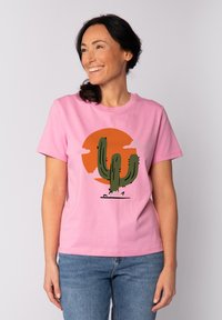 Roze katoenen T-shirt met een groene cactusafbeelding tegen een oranje zon en lichte wolken, met korte mouwen en een ronde hals.