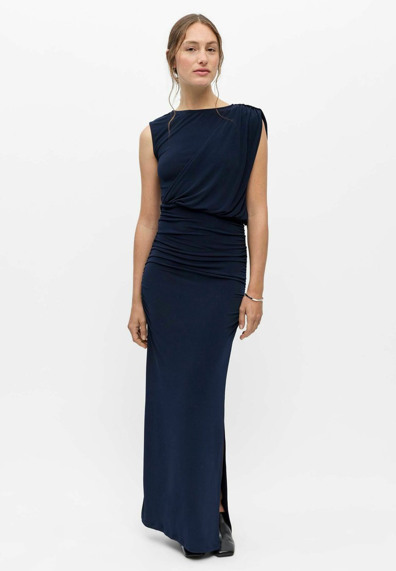 Femme portant une robe de soirée bleu marine sans manches froncée avec un décolleté asymétrique et des bottines noires, debout devant un fond blanc.
