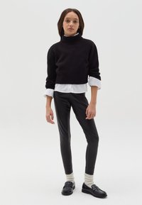 Un suéter negro de punto con cuello alto recortado sobre una camisa blanca de cuello, combinado con pantalones de cuero negros ajustados y mocasines negros con calcetines crema.