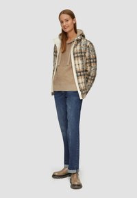 Geruite ritsjack met witte biezen, beige fleece hoodie, lichtbruine trui, blauwe spijkerbroek en bruine enkellaarzen met een dikke zool.