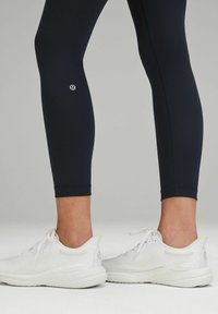 Navy leggings with a smooth texture feature a logo on the calf, paired with white athletic shoes with a textured upper and rounded sole.