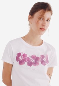 Valkoinen lyhythihainen t-paita, jossa on vaaleanpunaisia hibiskuskukkia ja teksti "HAWAIIAN LOVE" kuvan alla.