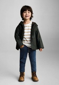Copil într-o jachetă verde închis peste o cămașă albă cu dungi galbene, blugi și cizme maro. Jacheta are glugă și nasturi.