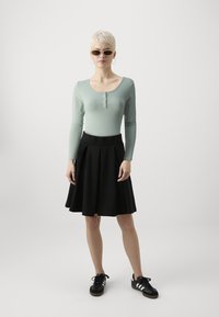 Lys grønn langermet bodysuit med knapper, svart plissert skjørt og svarte joggesko med hvite striper, komplementert av solbriller.