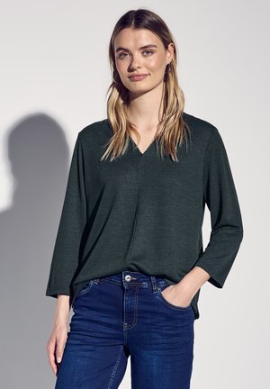 Top lungo verde scuro con scollatura a V, realizzato in tessuto morbido. Abbinato a jeans blu scuro, con una vestibilità comoda.
