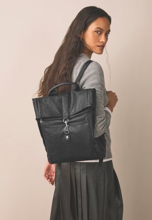 Kvinde med langt mørkt hår, iført en grå sweater og sort nederdel, bærer en sort læderrygsæk med metal spænde over den ene skulder.