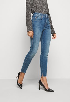 Frau trägt mittelblaue Skinny-Jeans mit Knöchelschlitzen, ein schwarz-weiß gemustertes Langarmshirt und schwarze spitze High Heels.