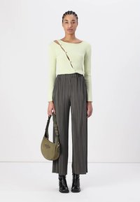 Femme portant un pull côtelé vert clair avec des boutons diagonaux, un pantalon large plissé noir, des bottes noires, et tenant un sac à épaule vert olive.