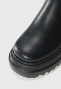 Close-up of black leather boot toe with thick rubber sole and textured grip pattern on white background.