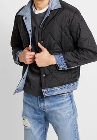 Svart vadderad jacka med denimdetaljer, med en ljusblå krage, tryckknappar och ribbade muddar. Bärs över en mörk sweatshirt.