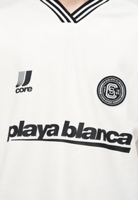 Λευκό πουκάμισο με μαύρη ριγωτή λαιμόκοψη σε σχήμα V, μαύρο κείμενο «playa blanca», λογότυπο Core αριστερά και στρογγυλή ετικέτα Athletic Club στο δεξί στήθος.