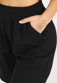 Svarta, strukturerade shorts med en elastisk midja och sidofickor. Tyget har ett subtilt mönster. En hand vilar i fickan och visar upp en tatuering.