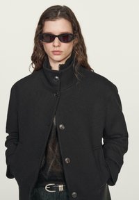 Cappotto nero in misto lana con colletto alto e cinque bottoni, abbinato a occhiali da sole neri slim. Il modello ha capelli mossi e uno strato sotto testurizzato.