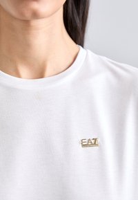 Tricou din bumbac alb, cu un mic patch cu logo-ul auriu EA7 pe partea stângă a pieptului. Gât rotund cu o textură fină.