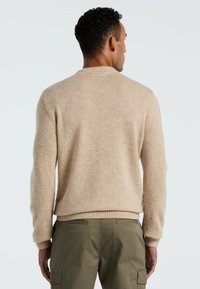 Man in een beige gebreide trui en olijfgroene cargo broek, van achteren gezien tegen een lichte, neutrale achtergrond.