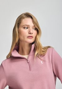 Ljusrosa zip-up sweatshirts med hög krage, prydd med en liten vit logotyp på vänster sida och en slät, mjuk textur.