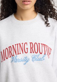 Grijze fleece sweatshirt met "MORNING ROUTINE" in het rood en "Varsity Club" in het blauw, met een ronde hals en een zachte textuur.