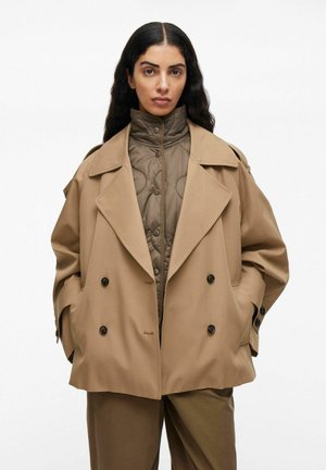 Beige dubbelzijdig jasje met grote revers en zwarte knopen, overlappend met een gewatteerde bruine binnenlaag. Gladde textuur en losse pasvorm.