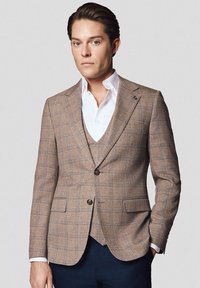 Brązowa marynarka w kratę wykonana z teksturowanej tkaniny z niebieskimi akcentami, z zapięciem na jeden guzik i dwiema przednimi kieszeniami. Noszona na białej koszuli.