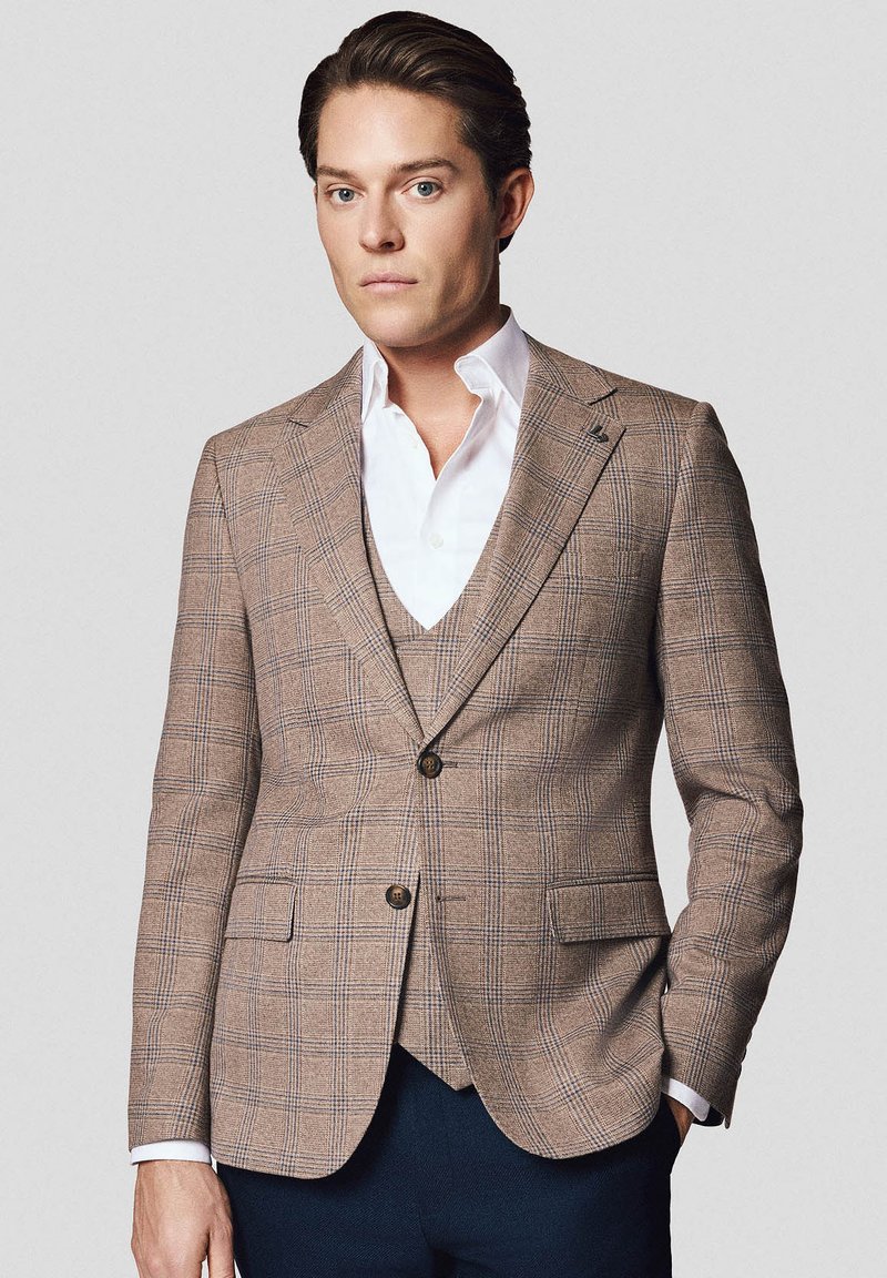 Brązowa marynarka w kratę wykonana z teksturowanej tkaniny z niebieskimi akcentami, z zapięciem na jeden guzik i dwiema przednimi kieszeniami. Noszona na białej koszuli.