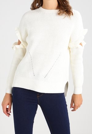 Femme portant un pull en maille blanc avec des détails découpés en volants sur les manches et un jean bleu foncé, debout sur un fond neutre.