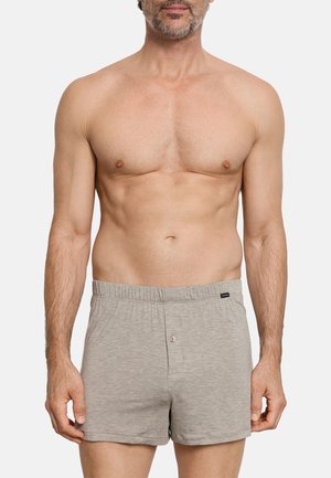 Uomo senza maglietta che indossa boxer in cotone grigio chiaro con chiusura a bottoni, in piedi contro uno sfondo bianco uniforme.