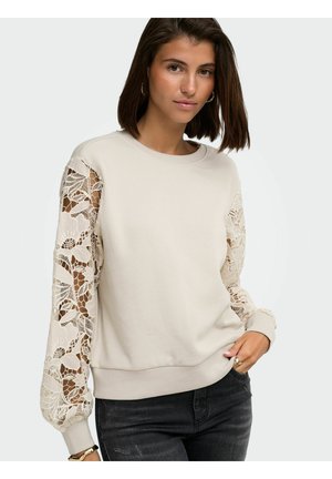 Donna che indossa una felpa beige con maniche lunghe in pizzo elaborato e jeans neri, in piedi davanti a uno sfondo chiaro uniforme.