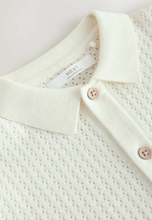 Polo in maglia color crema con motivo a rilievo, colletto classico e due bottoni con una leggera brillantezza. L'etichetta del marchio è visibile.