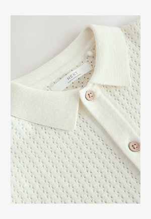 Cremefarbenes, gestricktes Polohemd mit strukturiertem Muster, klassischem Kragen und zwei Knopfverschlüssen mit dezentem Glanz. Das Markenlabel ist sichtbar.