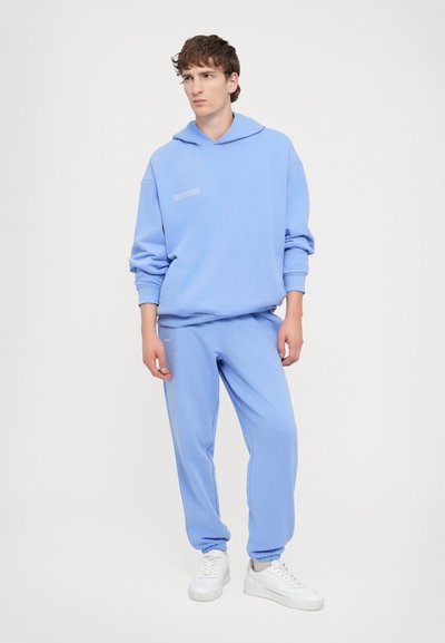 Light blue sweatshirt with a hood and loose fit, paired with matching joggers. Made of soft fabric; features subtle text detail. White sneakers complete the look.