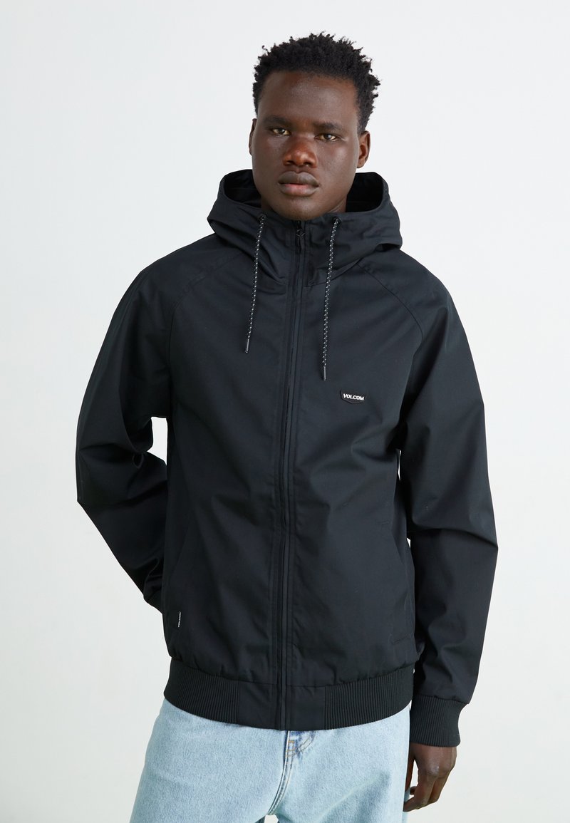 Chaqueta negra con cremallera y capucha, puños y cintura elásticos. Cuenta con una tela texturizada y un pequeño logo en el pecho.