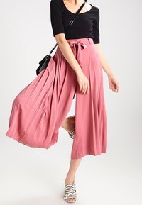 Rosa plissierte Culottes mit gebundenem Bund, kombiniert mit einem schwarzen, figurbetonten Oberteil und gestreiften Riemchenschuhen. Sanfter Stoff mit fließendem Design.