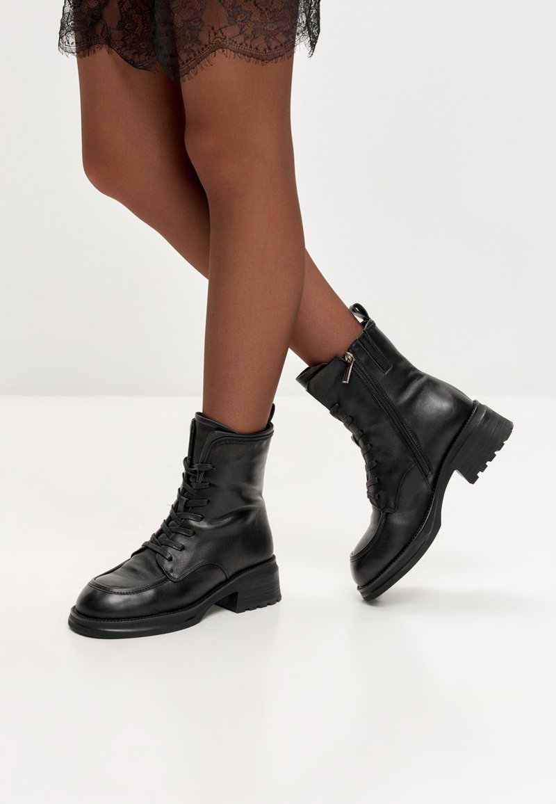 Nogi skrzyżowane, ubrane w przezroczyste brązowe rajstopy i czarne skórzane ankle boots z wkładką na zamek bocznym, stojące na białej podłodze.