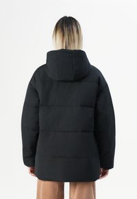 Veste matelassée noire avec capuche, présentant un design matelassé et une coupe décontractée. Texture lisse, manches longues et une coupe mi-longue.