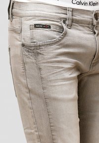 Světlé šedé džíny z denimu s úzkým střihem, které mají přední a zadní kapsy, jemné šití a zapínání na kovový knoflík.