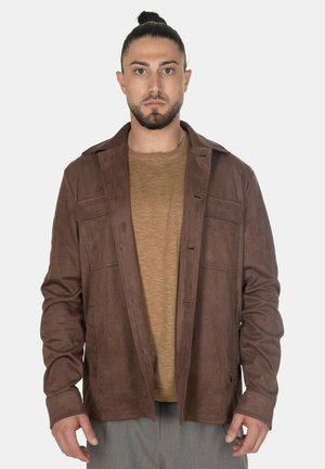 Uomo con capelli scuri raccolti e barba, indossa una giacca aperta in pelle scamosciata marrone su una camicia beige e pantaloni grigi, in piedi davanti a uno sfondo bianco.