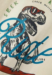 Camiseta blanca de algodón con un gráfico de un dinosaurio que lleva gafas de sol rojas, con letras en azul y negro, y "1986" impreso en la parte inferior.