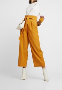 Weit geschnittene, senfgelbe Hosen mit einem Bindegürtel und glatter Textur, kombiniert mit einem weißen Pullover und cremefarbenen Knöchelstiefeln.