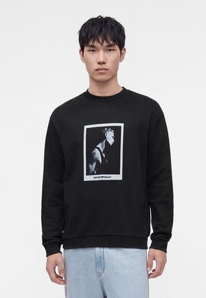 Jeune homme portant un sweat-shirt noir avec un motif blanc représentant une silhouette de profil et un jean bleu clair, debout devant un fond uni.