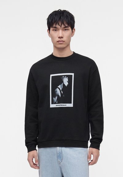 Jeune homme portant un sweat-shirt noir avec un motif blanc représentant une silhouette de profil et un jean bleu clair, debout devant un fond uni.