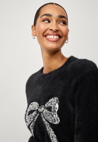 Zwarte fuzzy sweater met een grote, zilveren paillettenstrik op de voorkant. De halslijn is rond en de textuur lijkt zacht.