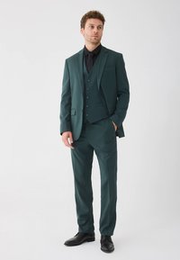 Tumši zaļš trīsdaļīgs uzvalks ar pieguļošu dizainu, teksturētu audumu un atbilstošām biksēm. Valkāts ar melnu kreklu un kaklasaiti. Melni apavi.