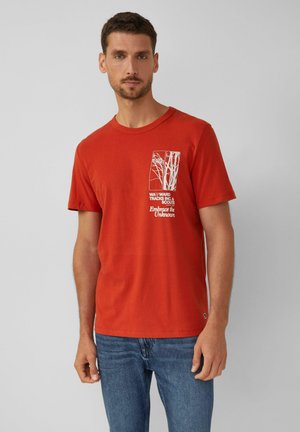 Man in een rode T-shirt met korte mouwen met een witte boomgrafiek en de tekst "Wayward Tracks Inc. & Scouts Omarm het Onbekende," blauwe spijkerbroek.